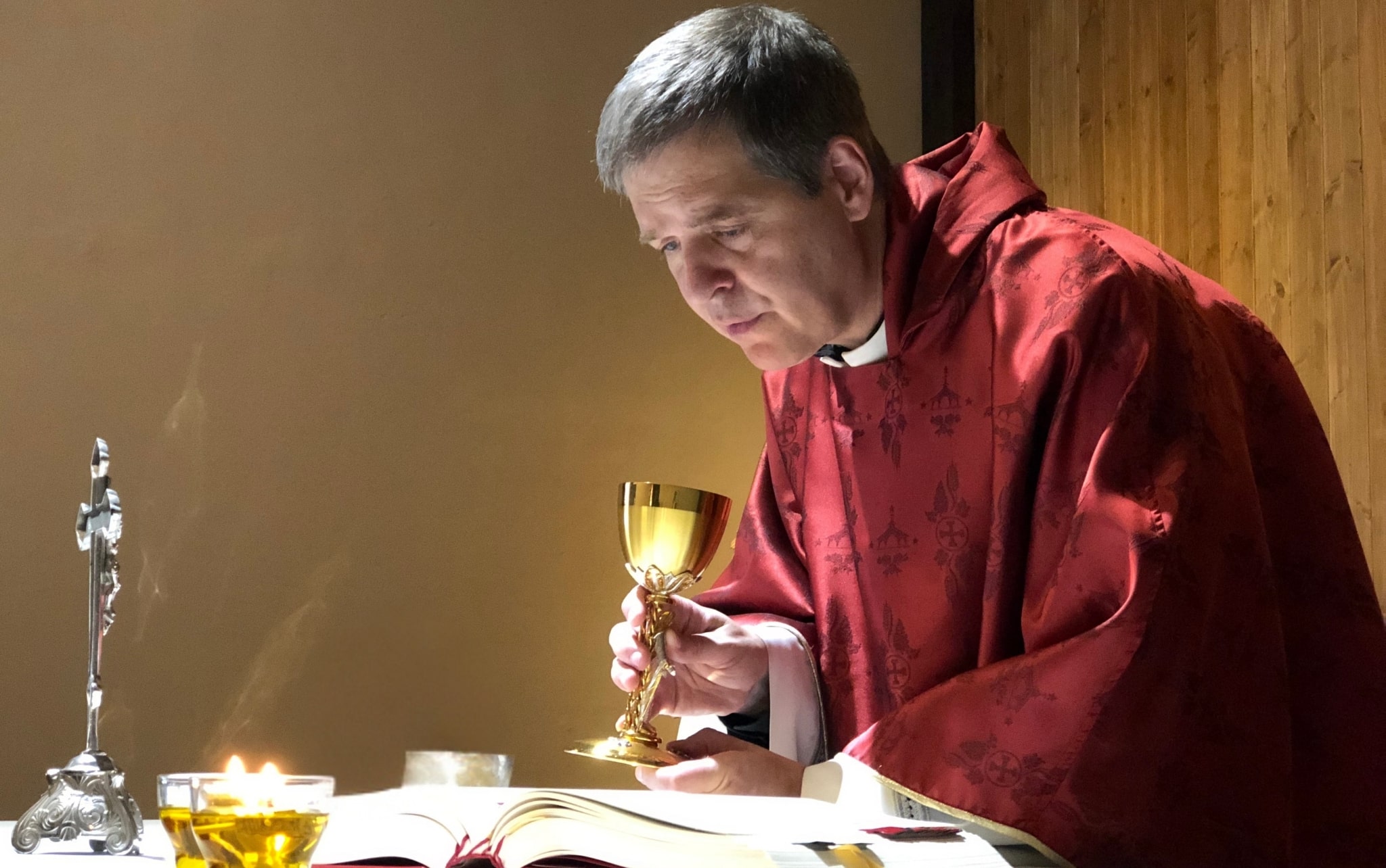 Fr. Henry in red chasuble reading from missal at altar with chalice and candles.