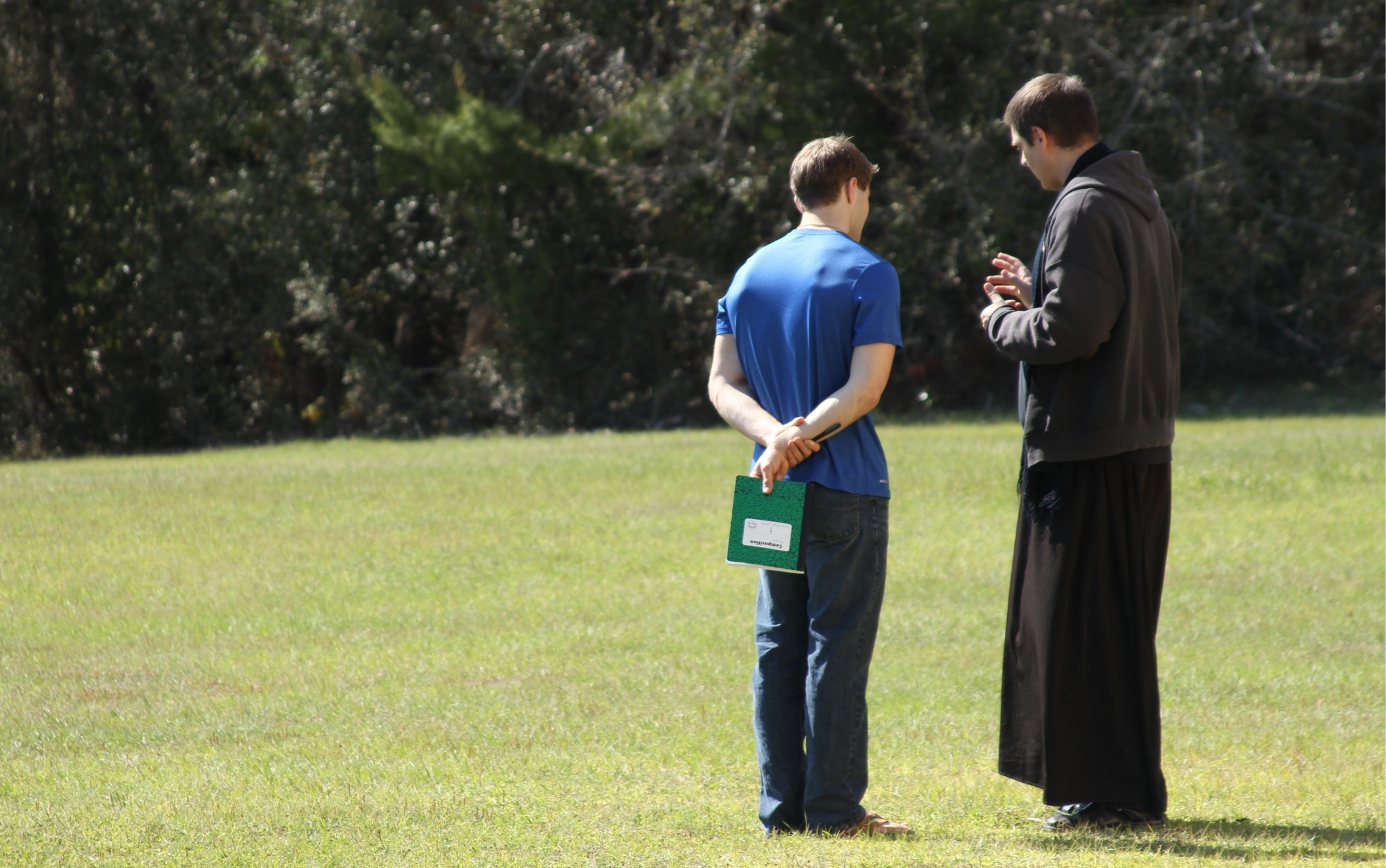 Young man in blue shirt holding green notebook in confession with Fr. Henry, who wears the black cassock. They are outside, on grass.